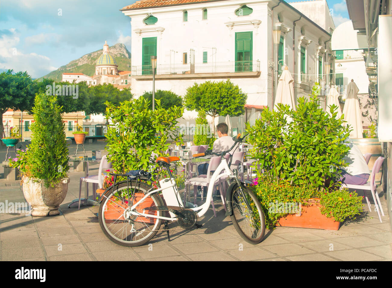 people having breakfast in morning at outdoor cafe with white bike in foreground and beautiful church in background on sunny summer day Stock Photo