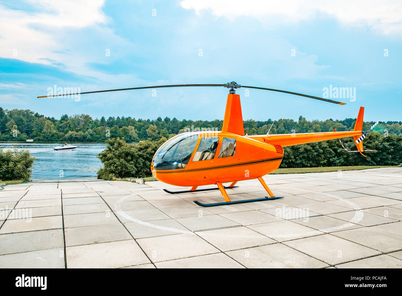 orange colored helicopter parked on a concrete slab under blue skies and water. Stock Photo