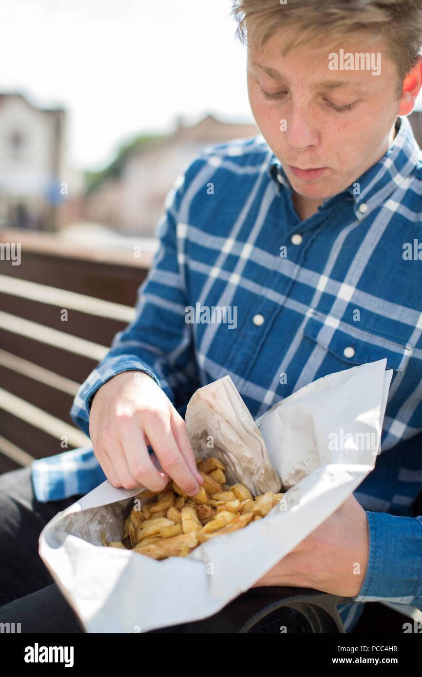 Teenage Boy Eating French Fries Sitting On Bench Outdoors Stock Photo