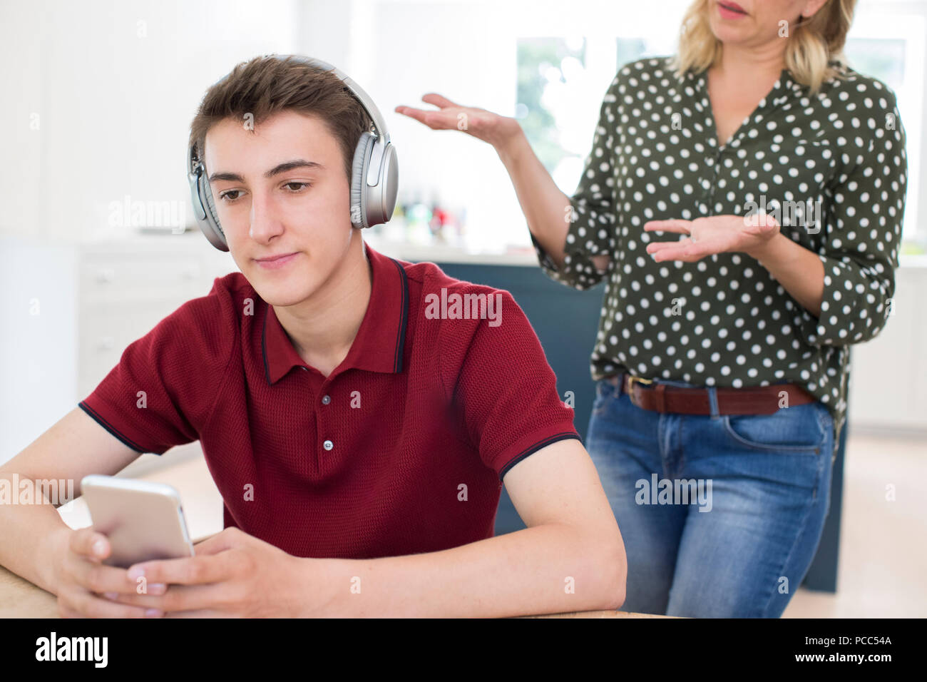 Teenage Boy Wearing Headphones And Using Mobile Phone Being Nagged By Mother At Home Stock Photo