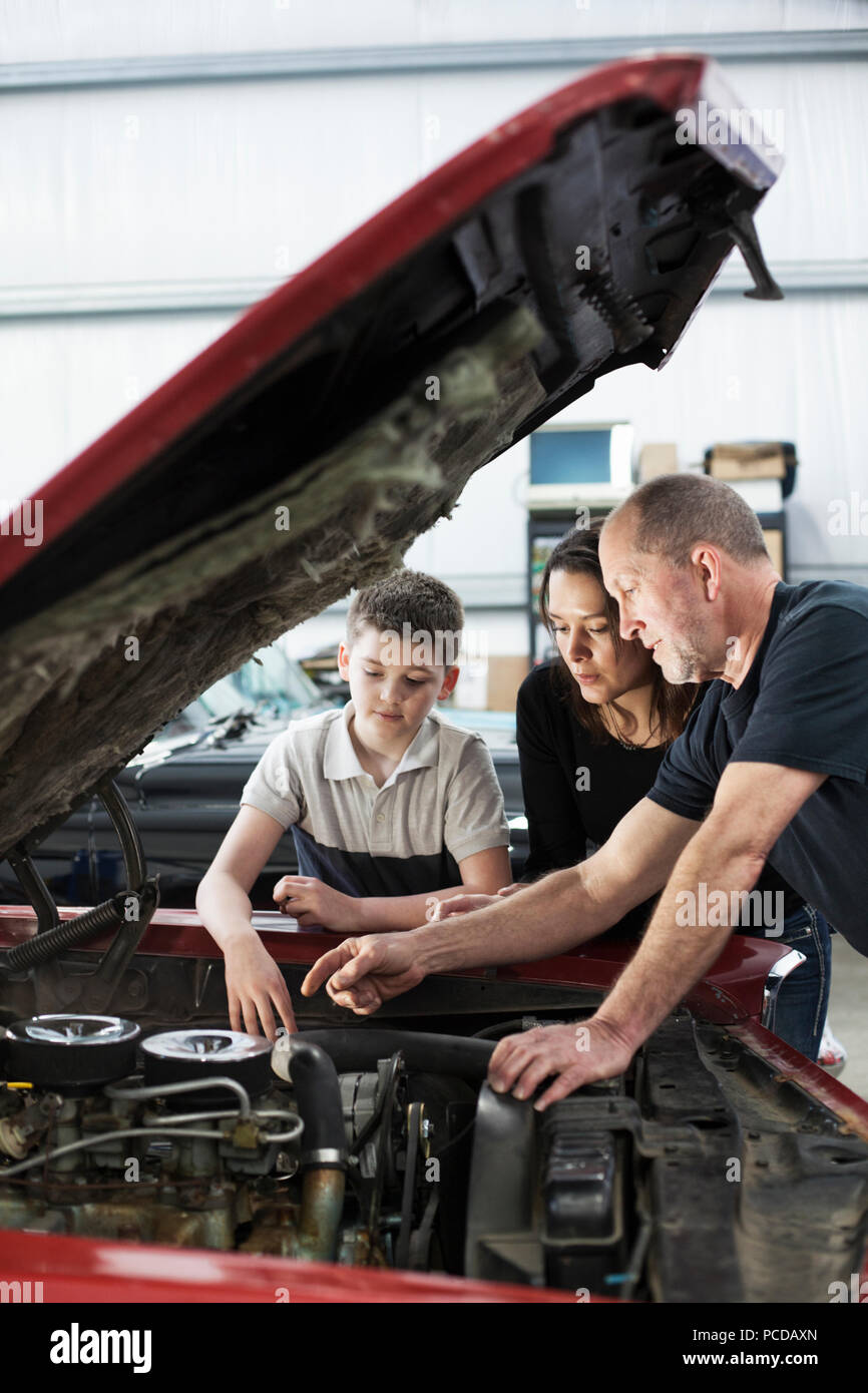 A senior caucasian male car mechanic talks to a caucsian mother and her son about a engine repair issue in a classic car repair shop. Stock Photo