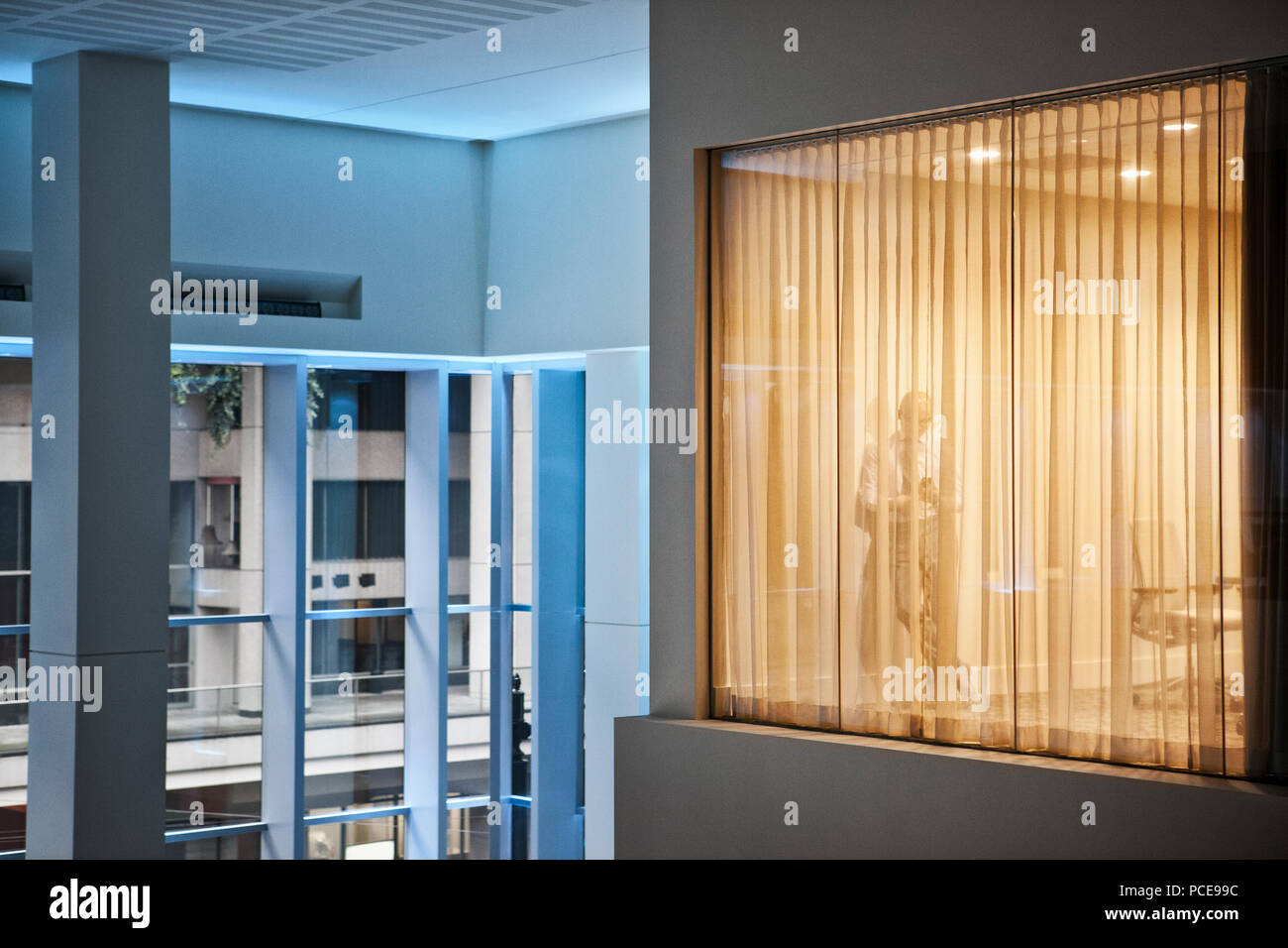 A man standing using h is phone in an office viewed from a building atrium. Stock Photo