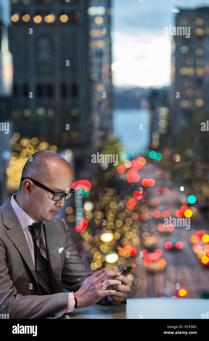 A side view of an asian businessman sitting at a table working on his phone in front of a window with a view to a city street at dusk. Stock Photo