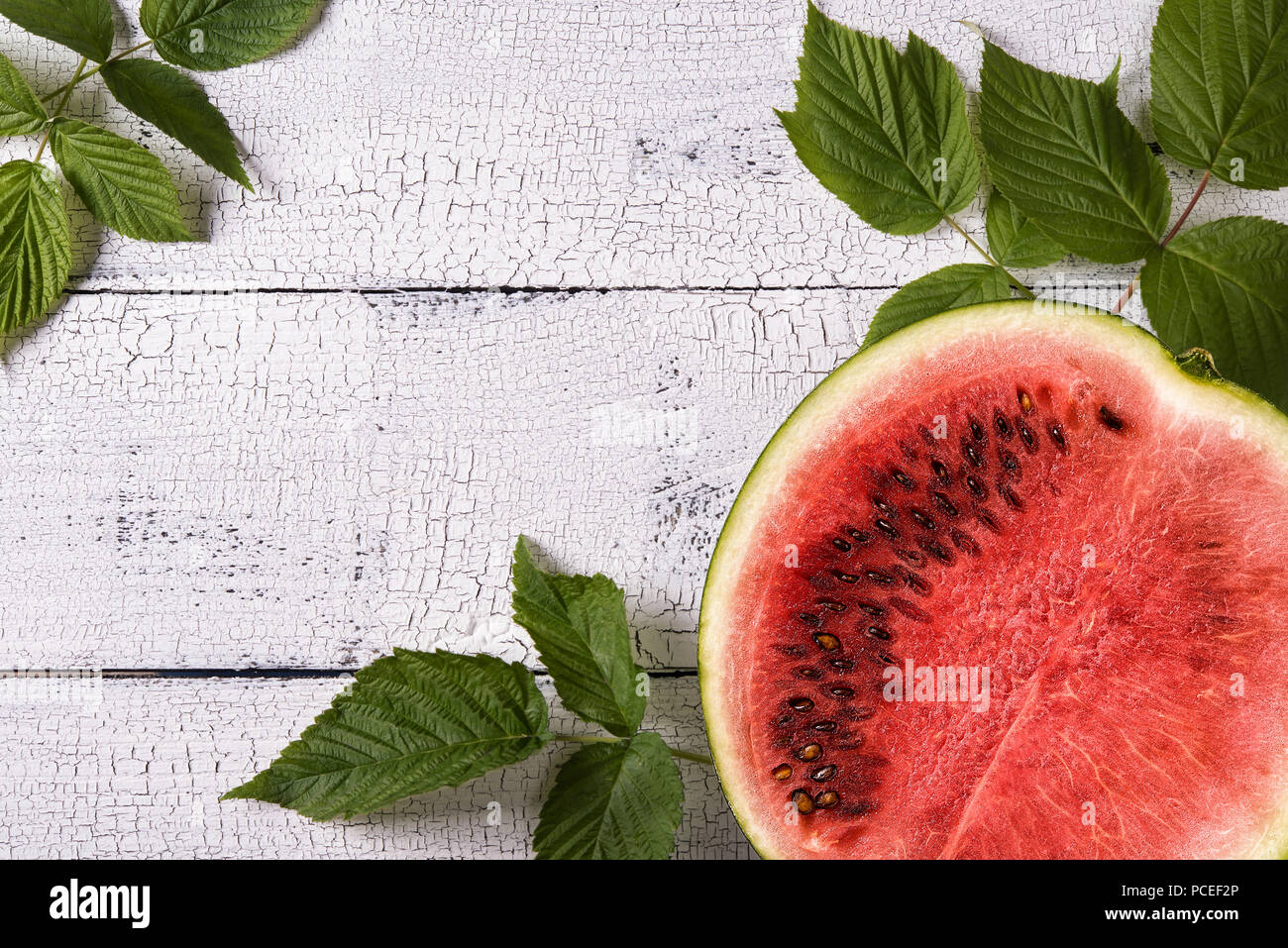 Cutted ripe red watermelon decorated with green leaves and on old white wooden table surface with cracks. Healthy eating and autumn harvest concepts.  Stock Photo