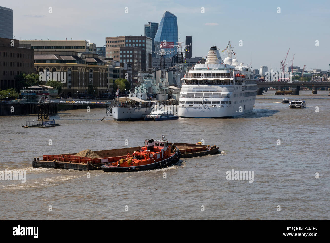 a tugboat pulling a barge and liter on the river thames in central london with hms belfast and the silversea cruise liner silver wind alongside. Stock Photo