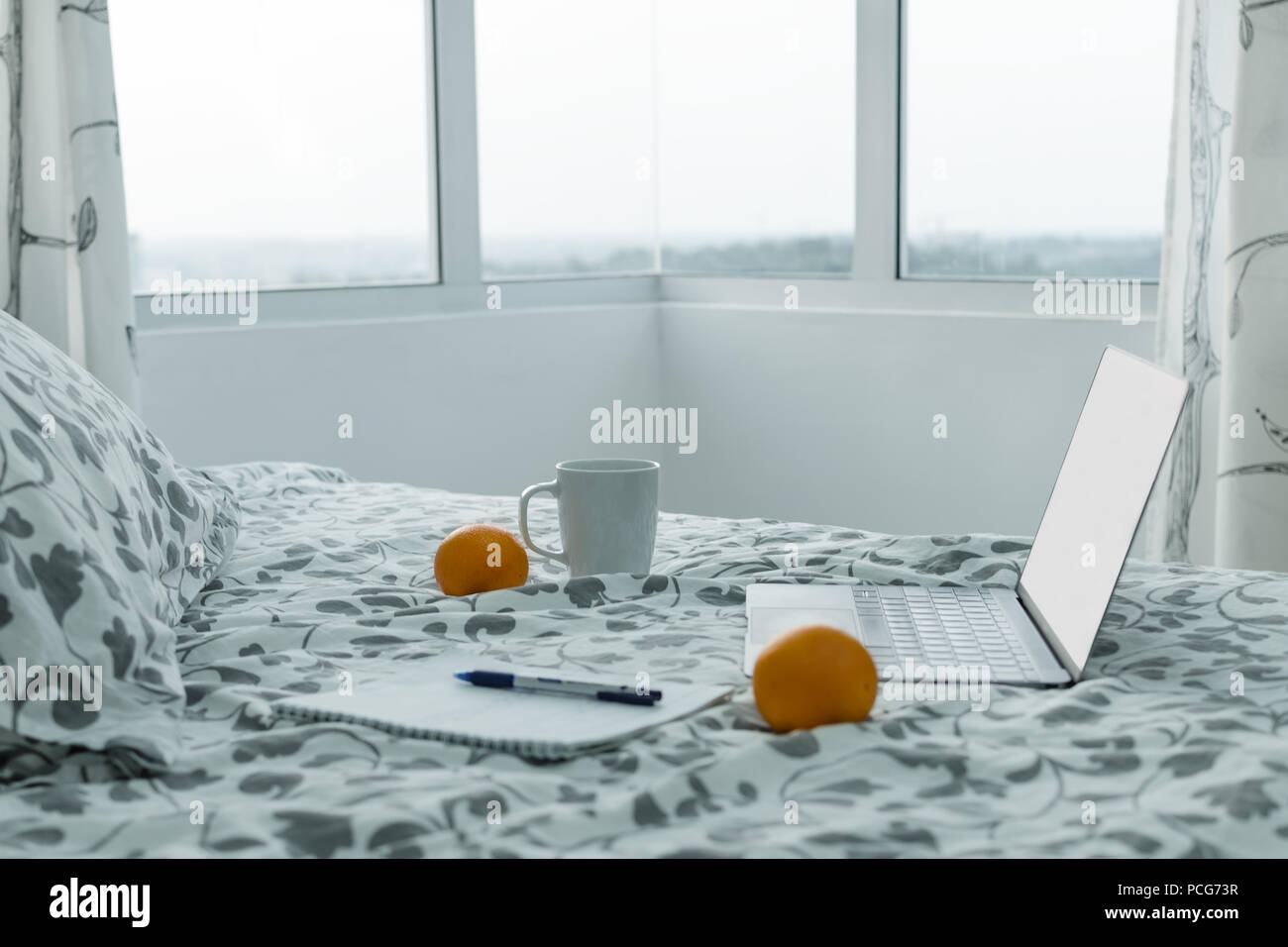 Morning in modern apartment - an open laptop on bed near window, next to cup of coffee, notebook, oranges. Concept of work at home Stock Photo