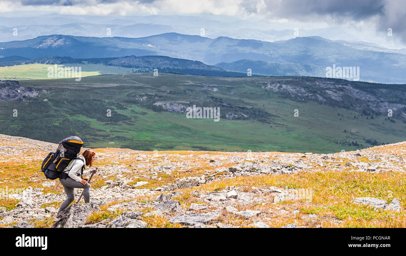 Atmospheric moment in mountains. Hiking woman with backpack traveler on top of mountains. Stylish woman hiking, in the background a green forest, fiel Stock Photo