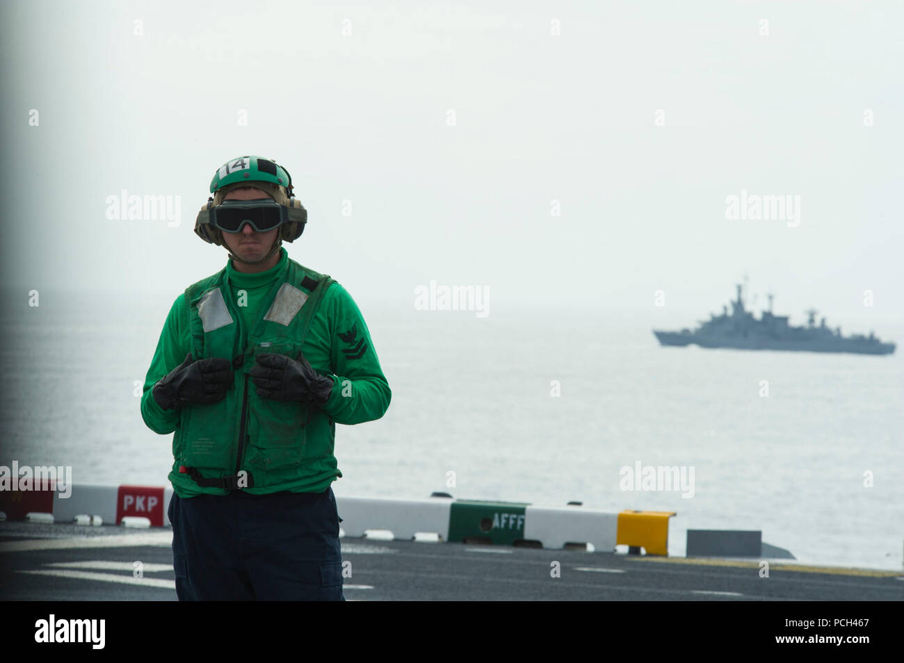 A U.S. Sailor assigned to amphibious assault ship USS America (LHA 6) waits on the flight deck as the Chilean navy frigate CNS Capitan Prat (FFGM 11), background, sails nearby during a bilateral exercise Aug. 27, 2014, in the Pacific Ocean. The America embarked on a mission to conduct training engagements with partner nations throughout the Americas before reporting to its new home port of San Diego. The ship was set to be ceremoniously commissioned Oct. 11, 2014. Stock Photo