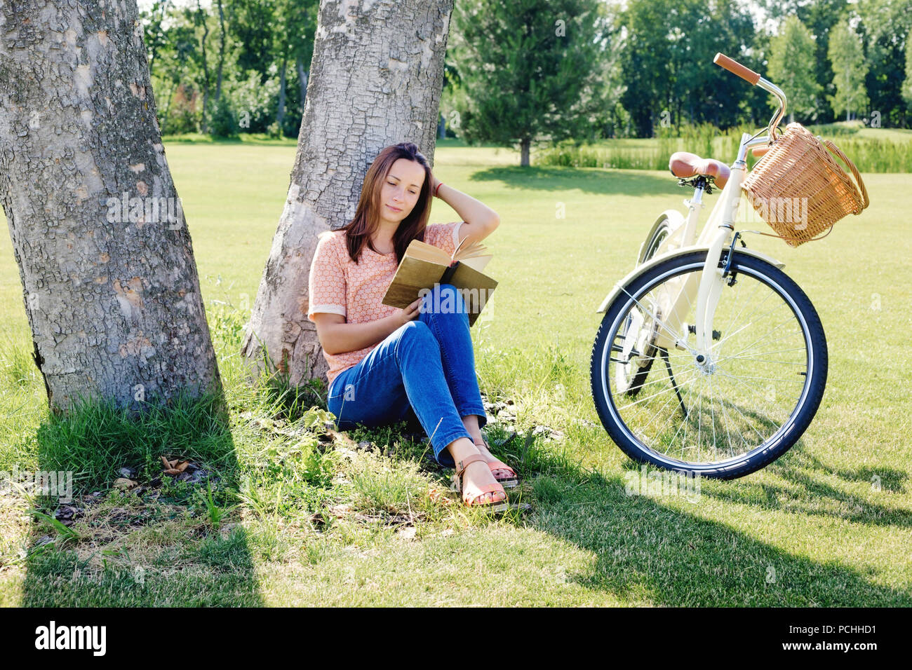 Image of young happy woman sitting under a tree with a book on a summer day Stock Photo