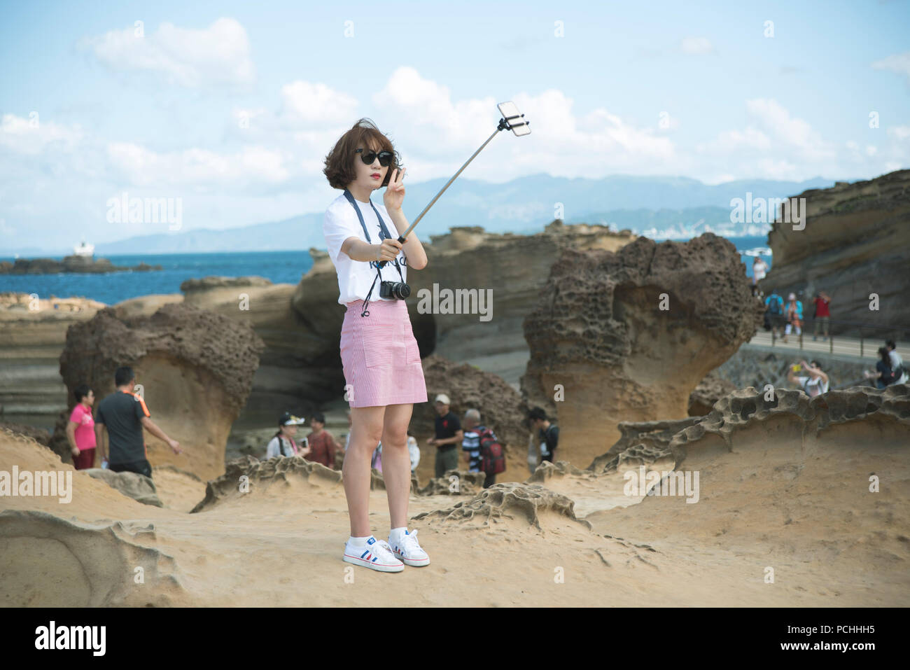 Chinese girl with sunglasses and a Bob haircut, wearing white sneakers, taking a selfie with a selfie stick in a geopark, with the sea in the back. Stock Photo