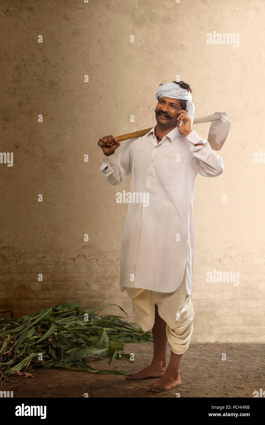 Indian farmer talking on mobile phone while carrying hoe on his shoulder Stock Photo