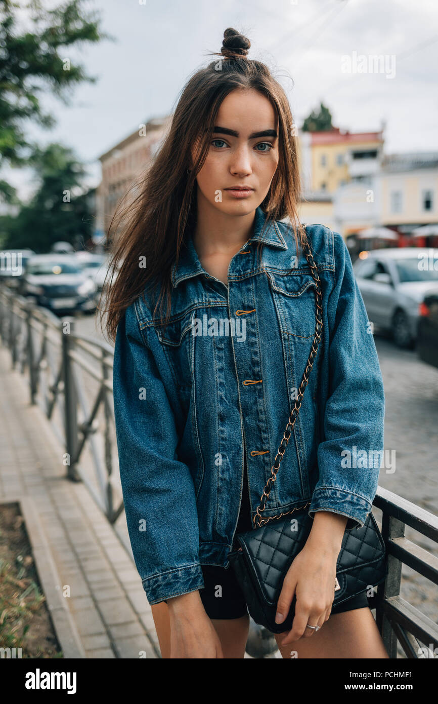 Young woman with long hair dressed in denim jacket standing on the street near road. Cute stylish teen girl posing in the city. Stock Photo