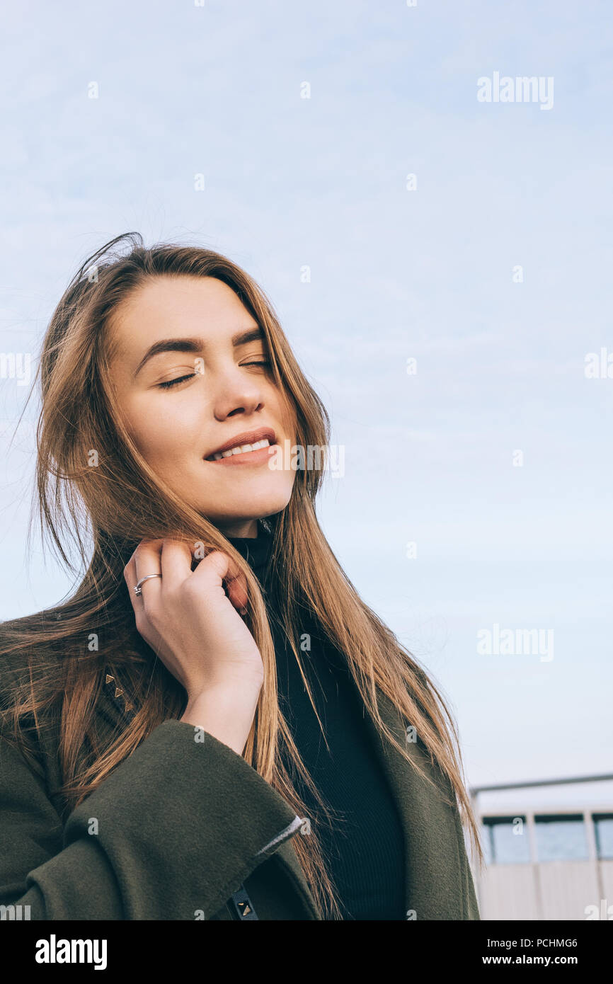 Young woman wearing stylish khaki coat and black turtleneck enjoying autumn day. Portrait of girl with long brown hair smiling and closing eyes outdoo Stock Photo
