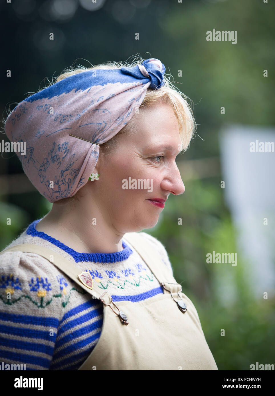 Close-up, side view portrait of isolated wartime housewife, wearing hand-knitted jumper, dungarees & head scarf at Black Country Museum 1940's event. Stock Photo