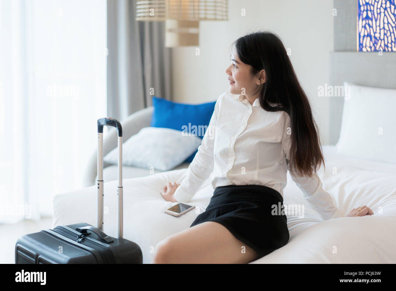 Smiling Asian business woman sitting on bed in hotel room. Business travel concept. Stock Photo