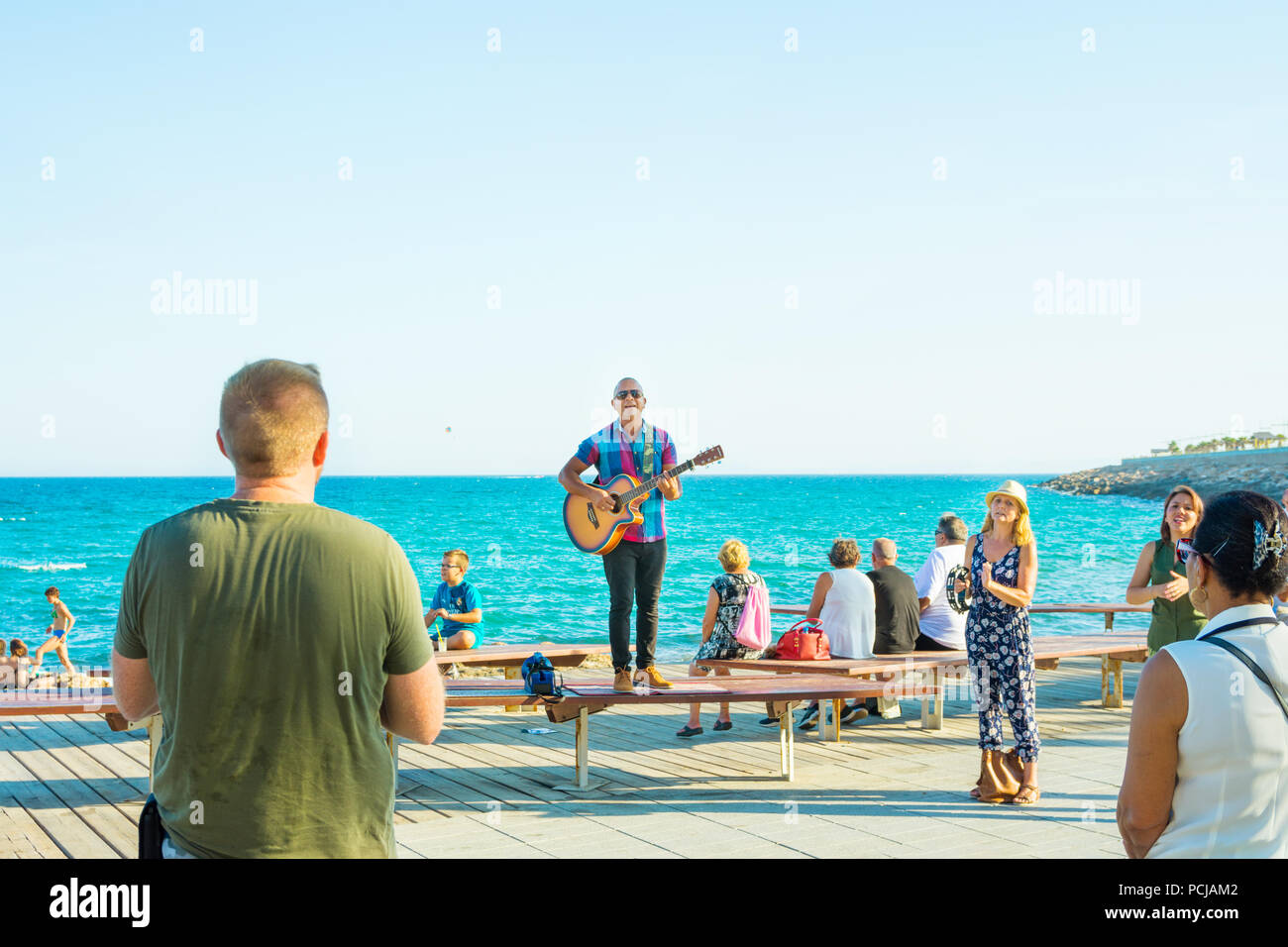 SPAIN, ALICANTE - JULY 28, 2018 Street band performing on seaside boardwalk on sunny summer day. Young man with guitarr singing audience applauding. B Stock Photo