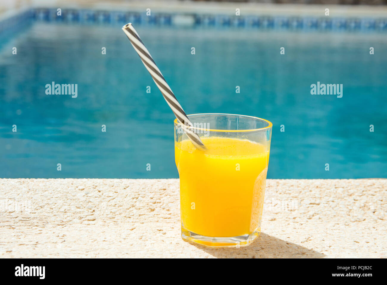 Glass of freshly pressed tropical fruits orange juice with striped straw standing on deck of swimming pool. Bright sunlight. Summer vacation relaxatio Stock Photo