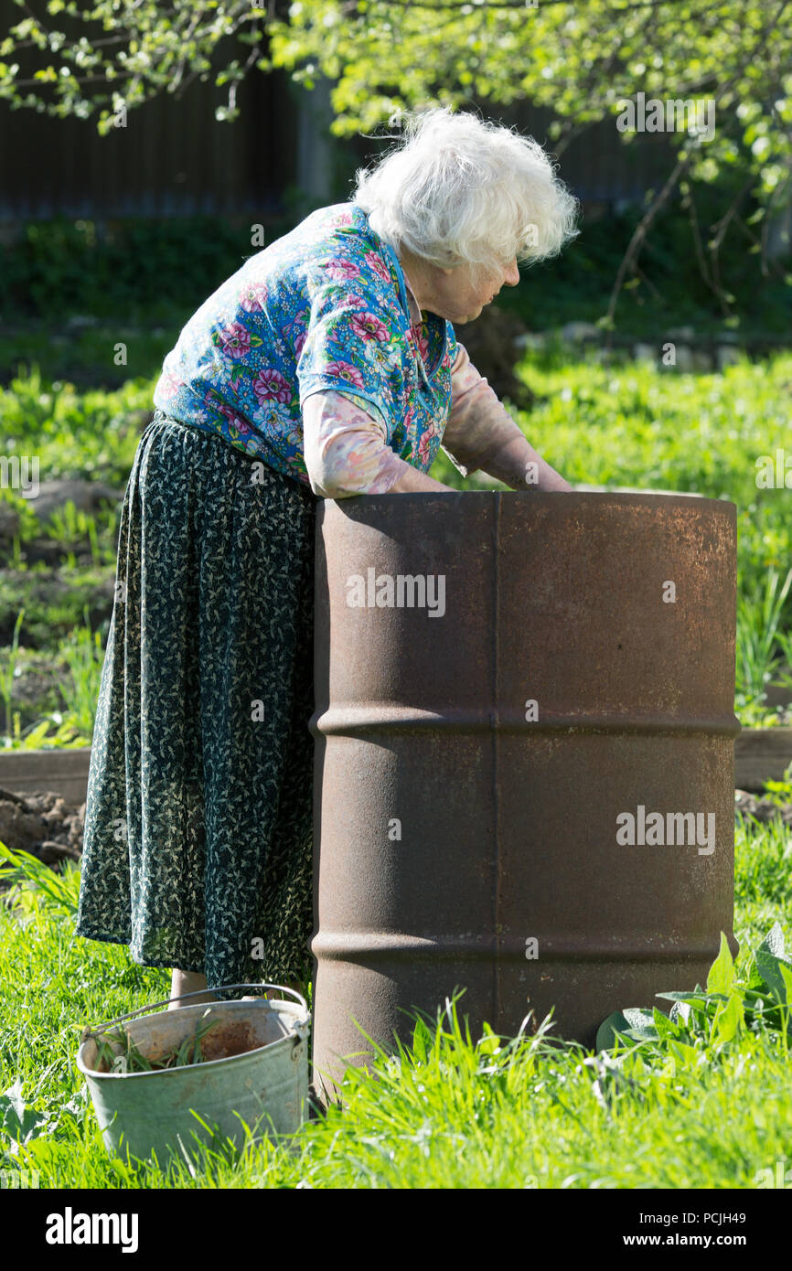 old woman in the garden at an iron barrel with water Stock Photo