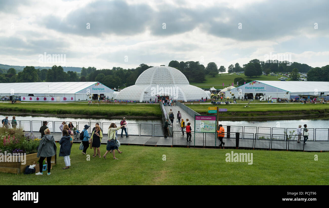 People at RHS Chatsworth Flower Show walking & crossing temporary river bridge leading to large marquees at rural showground - Derbyshire, England, UK Stock Photo