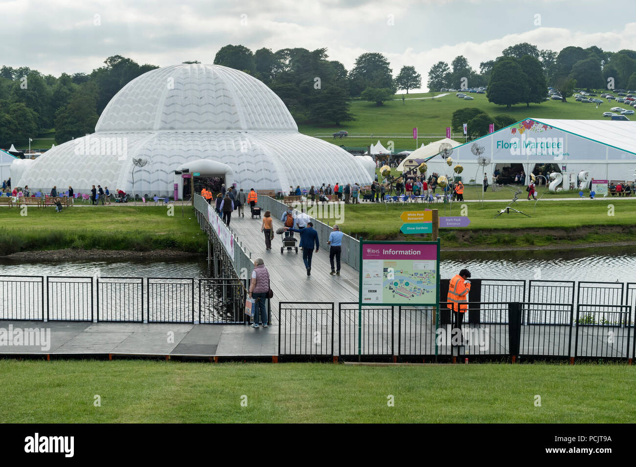 People at RHS Chatsworth Flower Show walking & crossing temporary river bridge leading to large marquees at busy showground - Derbyshire, England, UK. Stock Photo