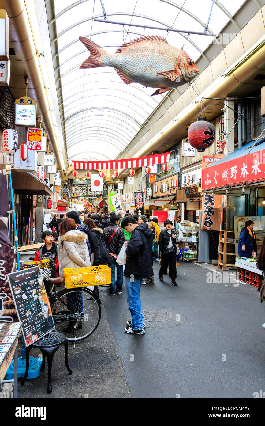 Kuromon Ichiba, Osaka's kitchen food market. View along arcade with shoppers and tourists walking through. Model fish hanging from roof. Wintertime. Stock Photo