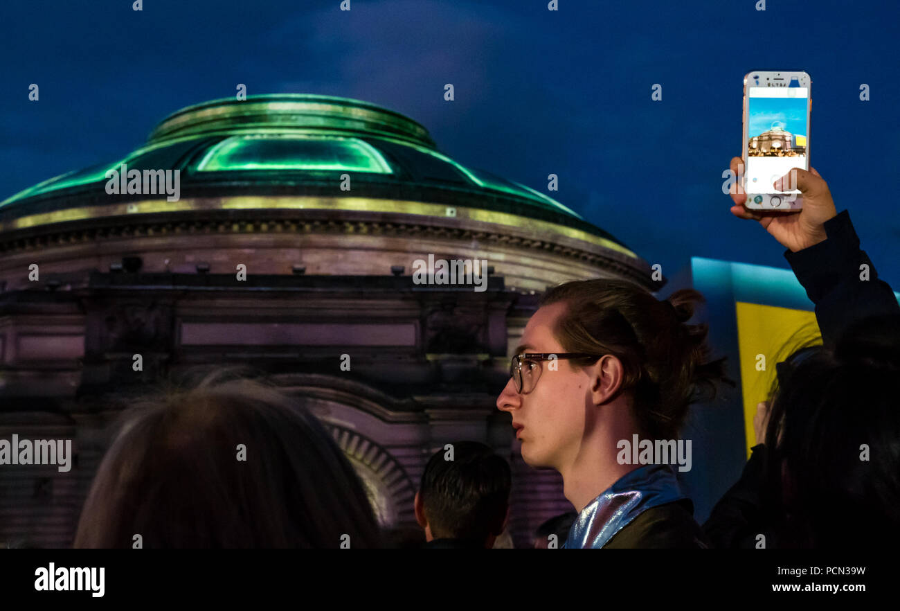 Edinburgh, UK. 3rd Aug 2018. Aberdeen Standard Investments sponsors the Edinburgh International Festival 2018 opening event Five Telegrams with a crowd in Festival Square. Celebrating Scotland’s Year of Young People and reflecting on the centenary of the end of the Great War with commissioned music by Anna Meredith and digital projection onto the Usher Hall by 59 Productions, the free outdoor event is Inspired by telegrams sent by young soldiers in 1918. One of the singers waiting to sing in the second Act called A Field Postcard while a person takes a photo on a mobile phone Stock Photo
