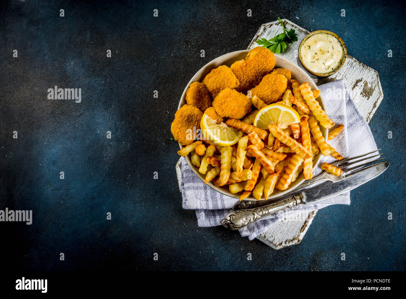 English traditional fast food, junk food, British dish Fish and chips with tartar sauce, lemon and greens, on a dark blue background, copy space above Stock Photo