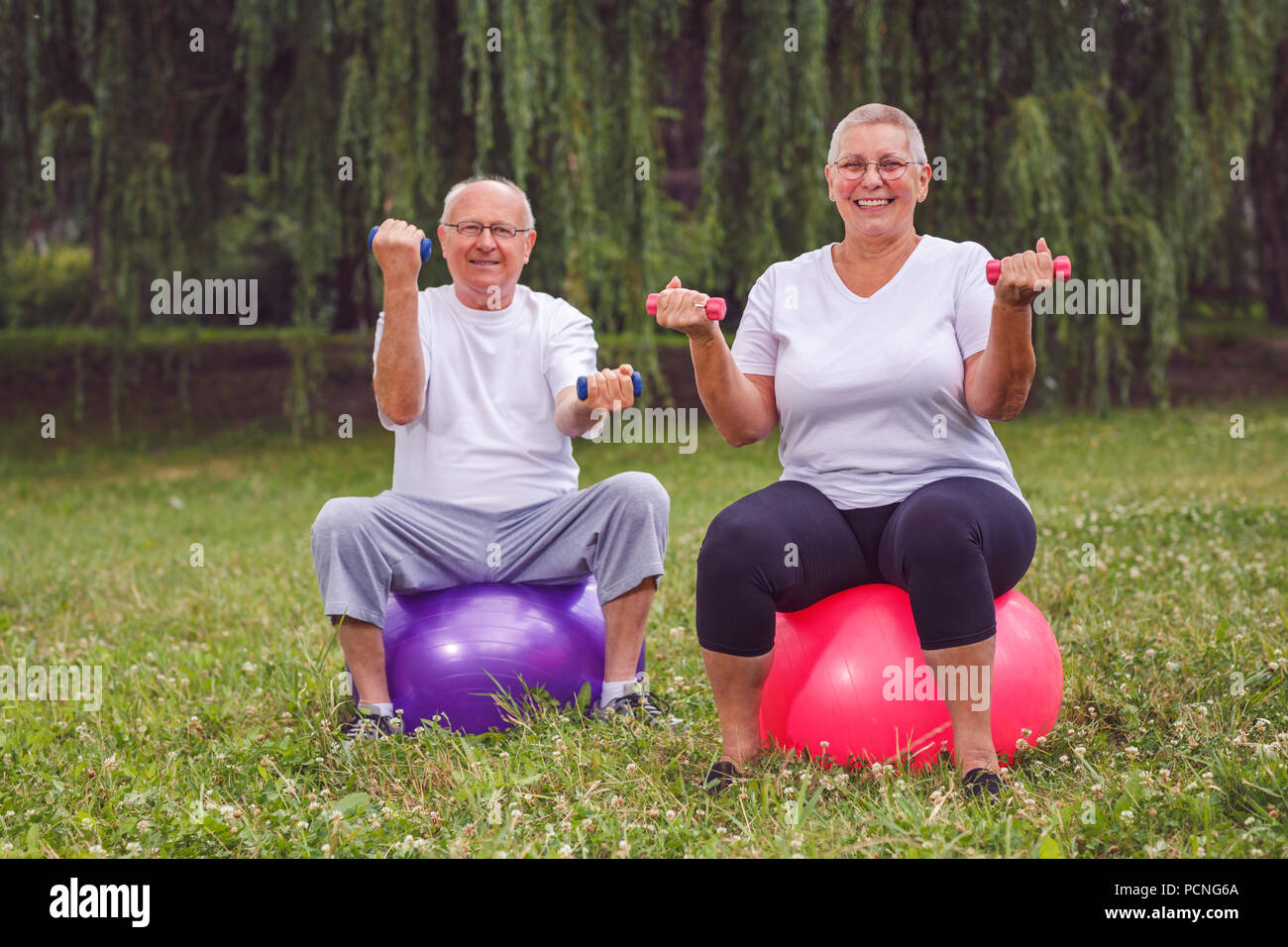 Smiling pensioner couple holding dumbbells while sitting on fitness ball in park Stock Photo