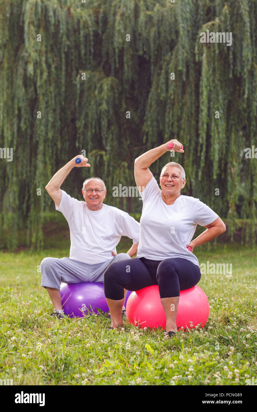 Smiling pensioner couple doing fitness exercises on fitness ball in park Stock Photo