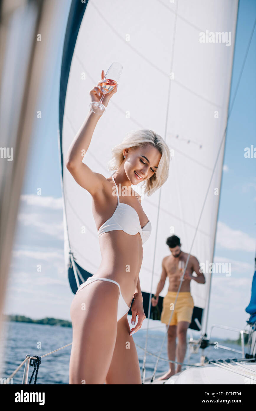 smiling woman in bikini holding champagne glass and her boyfriend walking behind on yacht Stock Photo