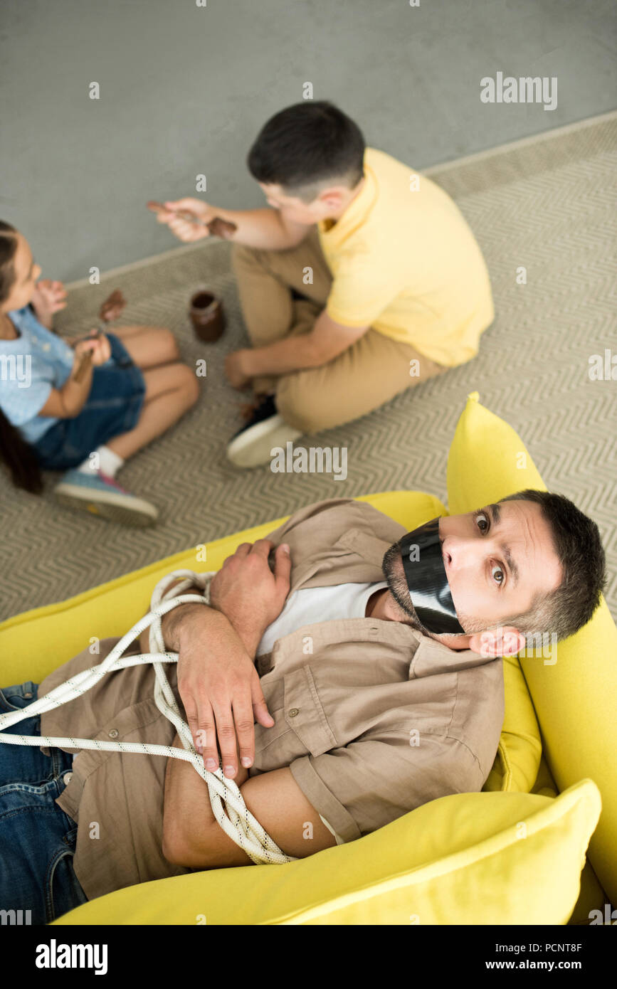 children eating chocolate and father lying tied on sofa and looking at  camera Stock Photo - Alamy