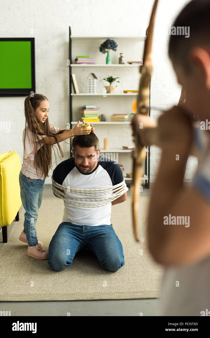 disobedient children playing with tied father and pretending shooting with toy bow at home Stock Photo