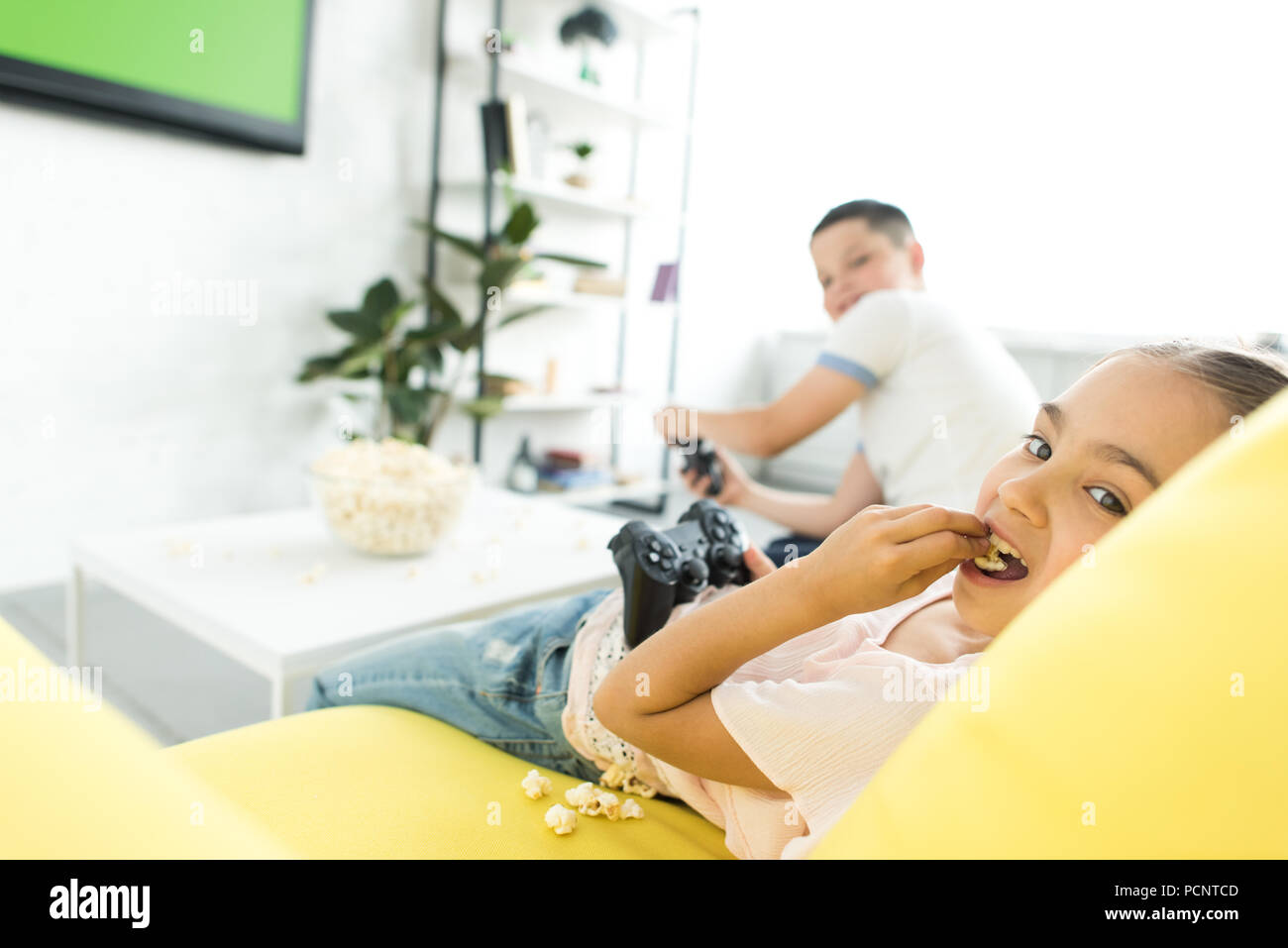 sister and brother playing video game and eating popcorn at home Stock Photo
