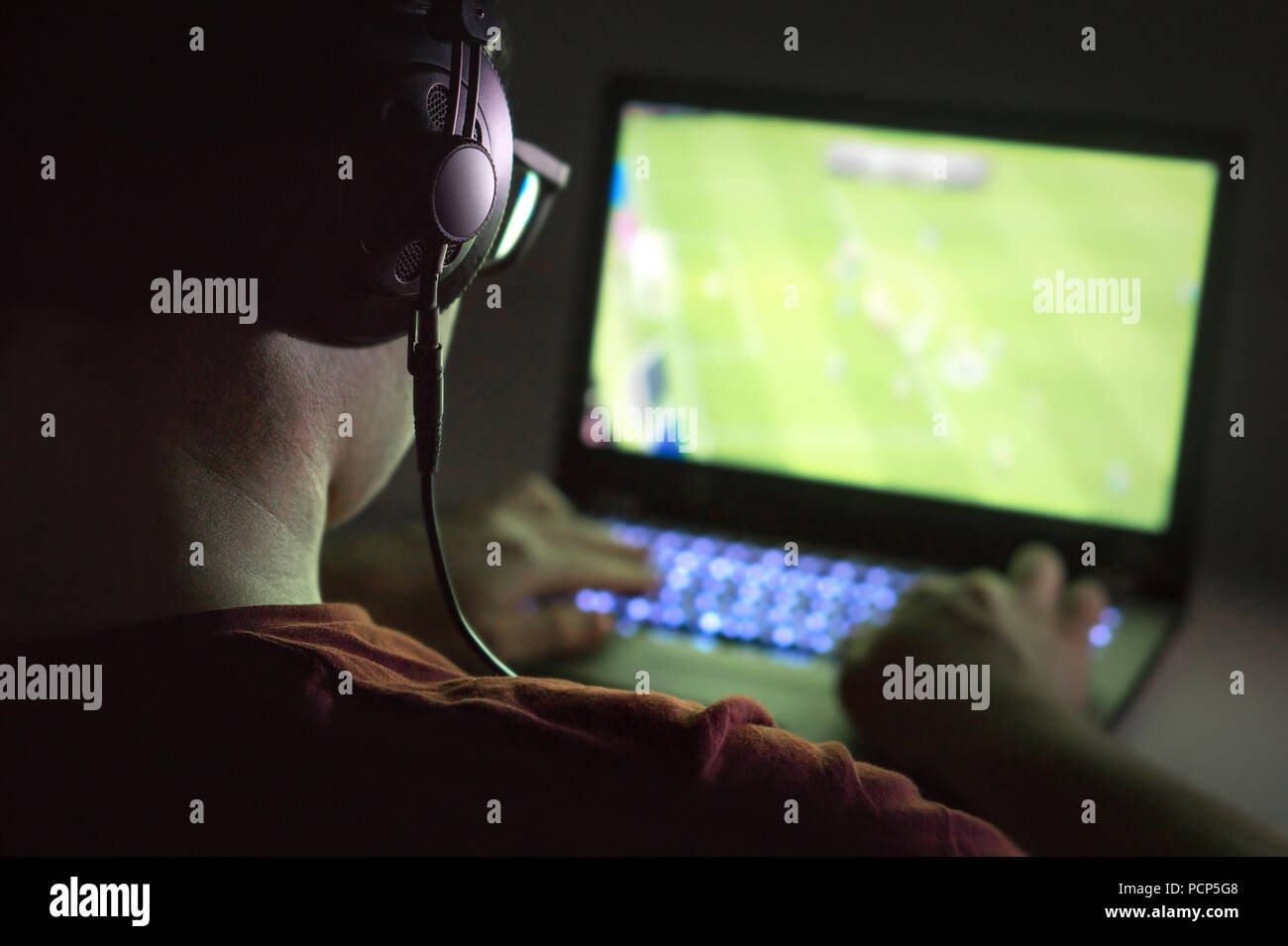 Playing video games with laptop. Young man plays online soccer or football on computer. Back view of gamer with headphones in dark or late at night. Stock Photo