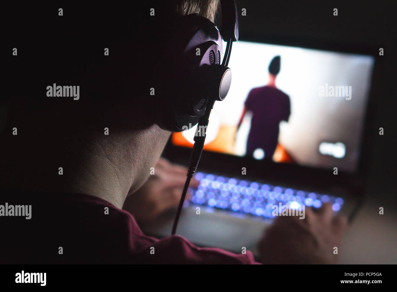 Playing video games with laptop. Young man plays action game on computer. Back view of gamer with headphones in dark or late at night. Stock Photo