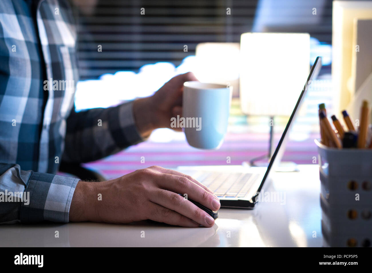 Working late at night and drinking coffee. Man using laptop computer and holding cup or mug in hand in home or office. Stock Photo