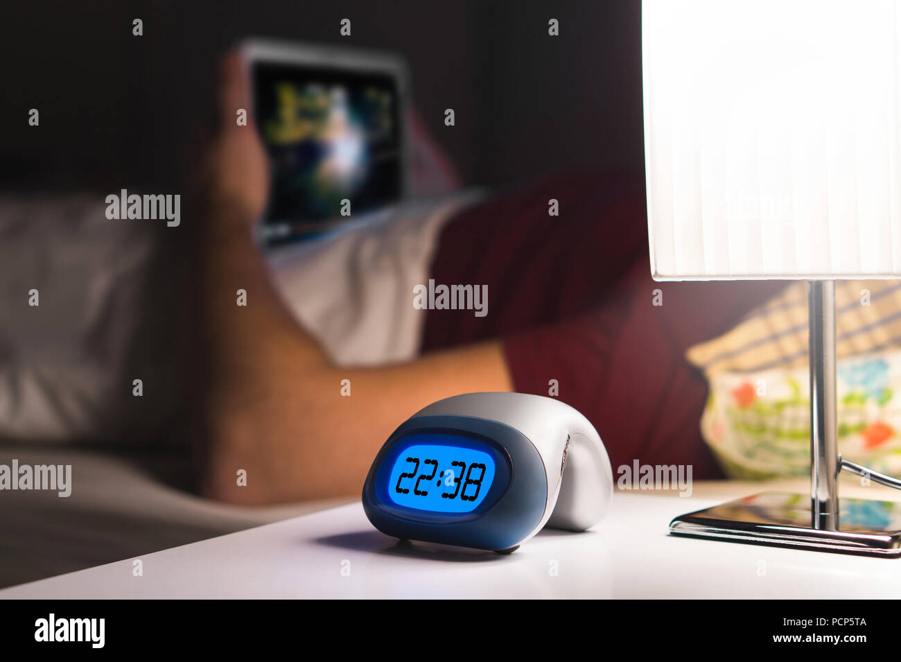 Man using tablet or watching movie or series stream online in bed before falling asleep. Digital alarm clock on nightstand in bedroom. Stock Photo
