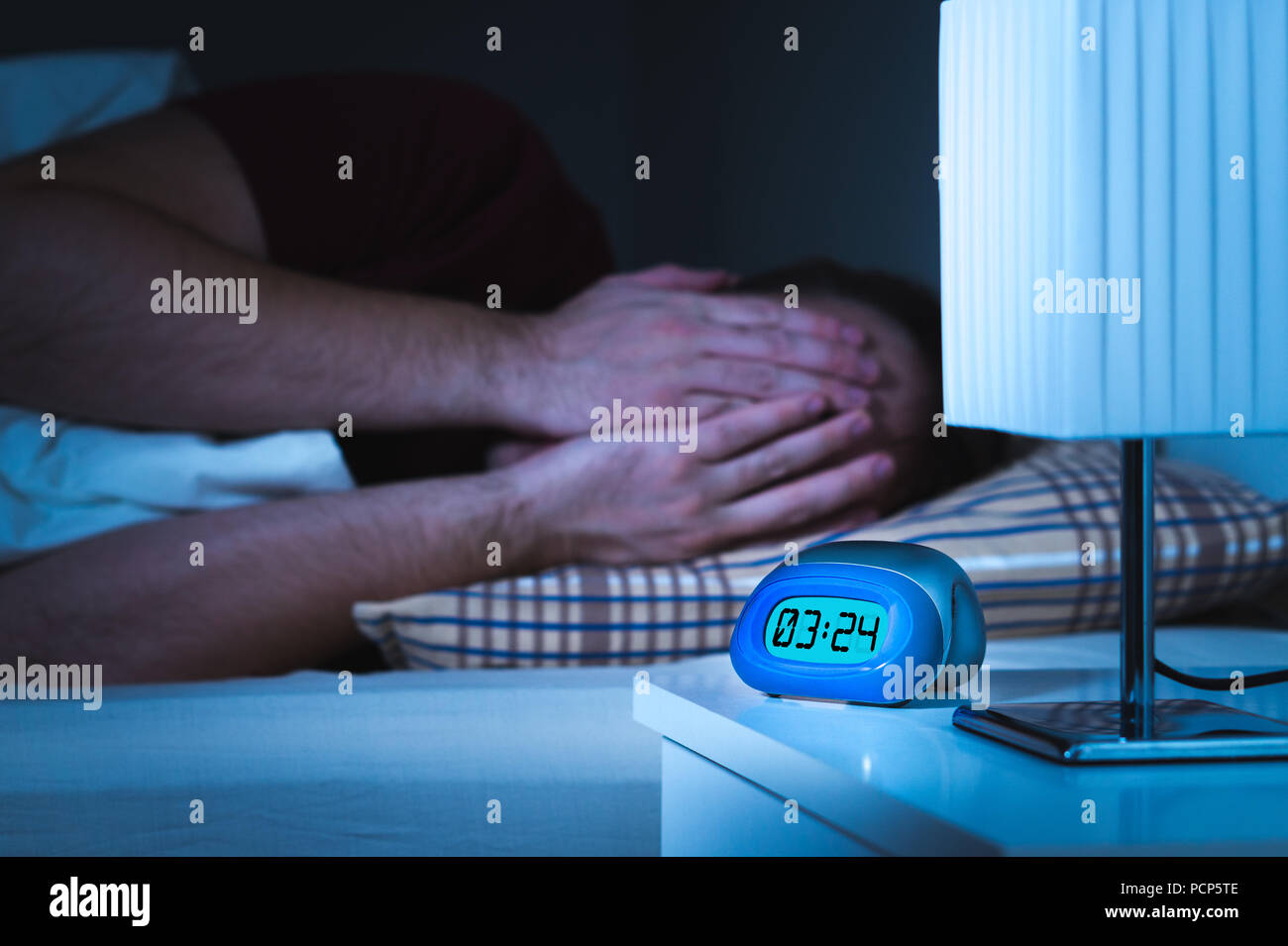 Seeing nightmares or bad dreams concept. Scared man covering face with hands in bed. Alarm clock on nightstand in bedroom. Stock Photo