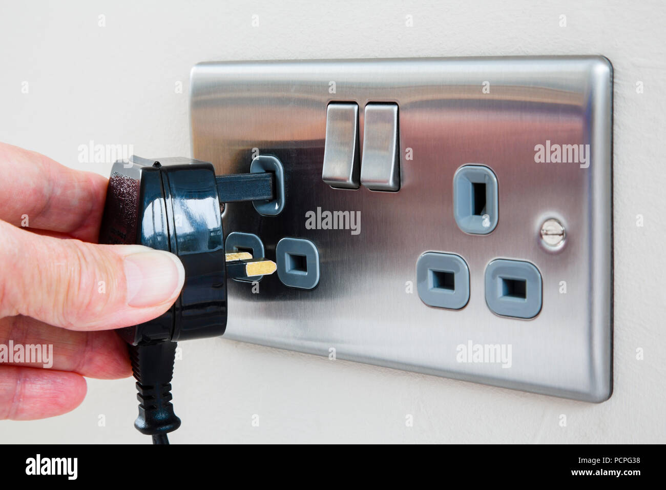 A person's hand holding an unplugged three pin plug and plugging it in to an electric wall socket switched off. England, UK, Britain Stock Photo