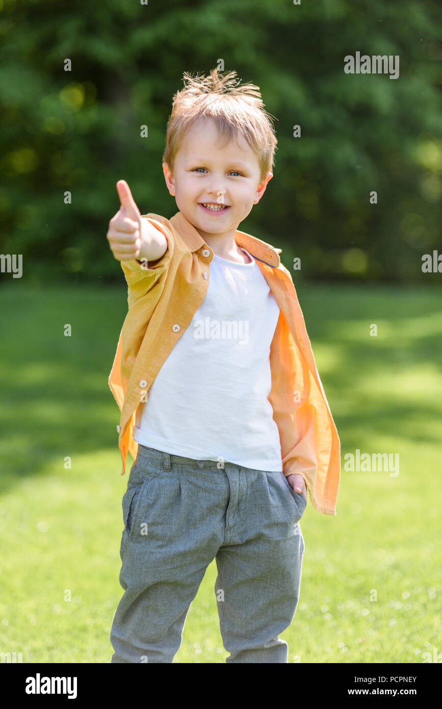 cute happy little boy showing thumb up and smiling at camera in park ...
