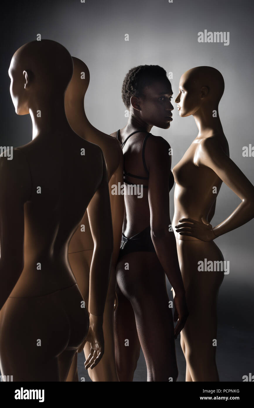 young african american model in bodysuit standing between dummies on black Stock Photo