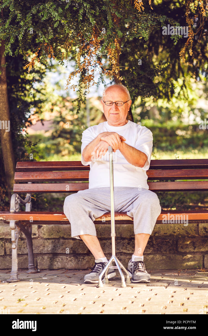 Sad pensioner man with his walking stick sitting on bench in the park Stock Photo