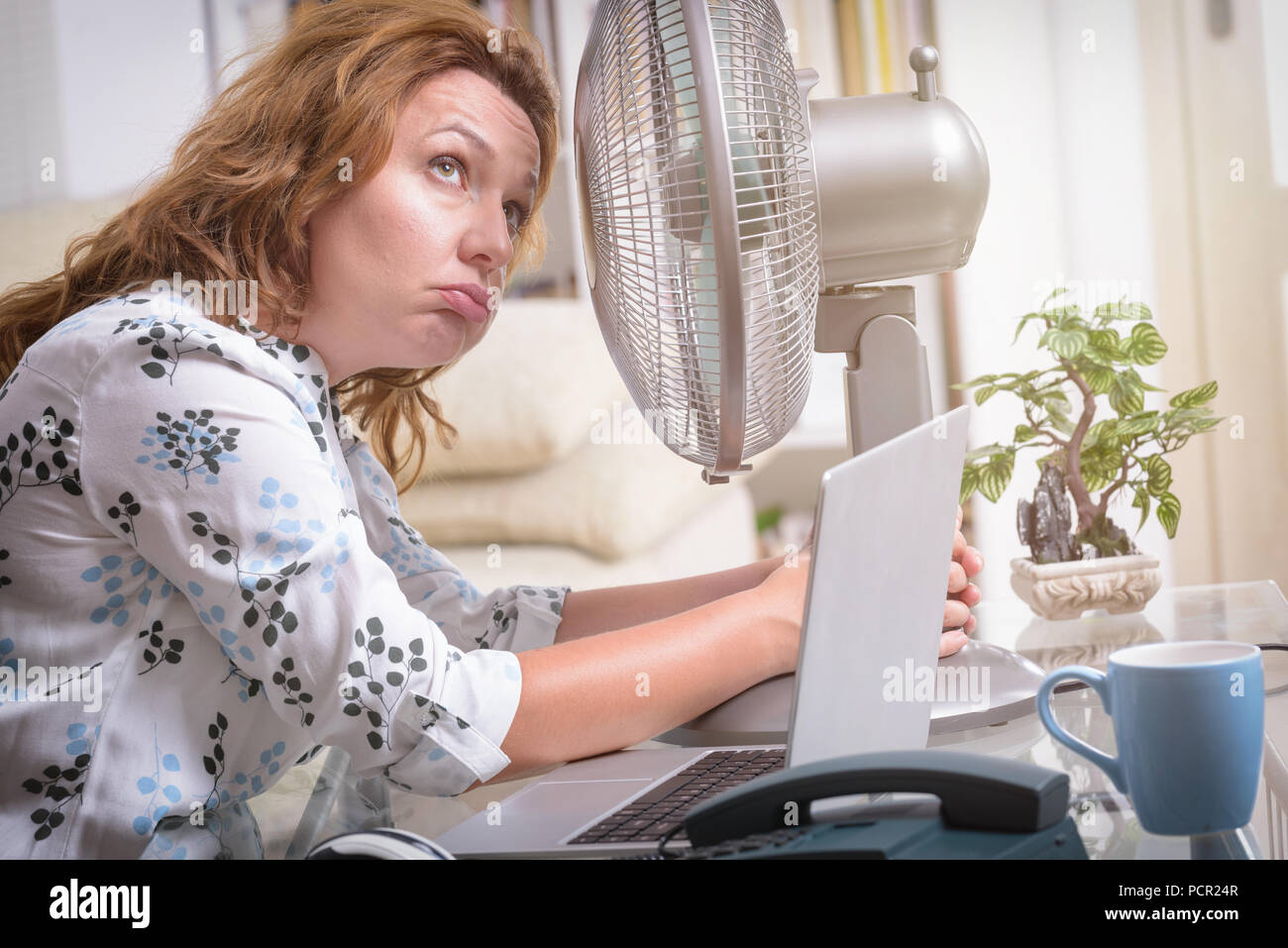 Woman suffers from heat while working in the office and tries to cool off by the fan Stock Photo