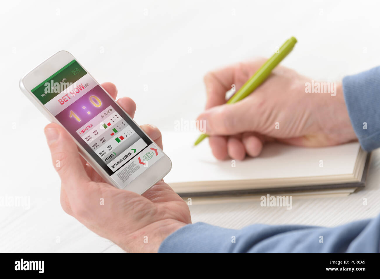 Man betting on sports, over shoulder view on hand with smartphone with scores and other hand taking notes Stock Photo