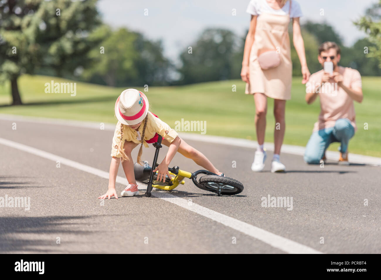 parents photographing little daughter falling from bicycle in park Stock Photo