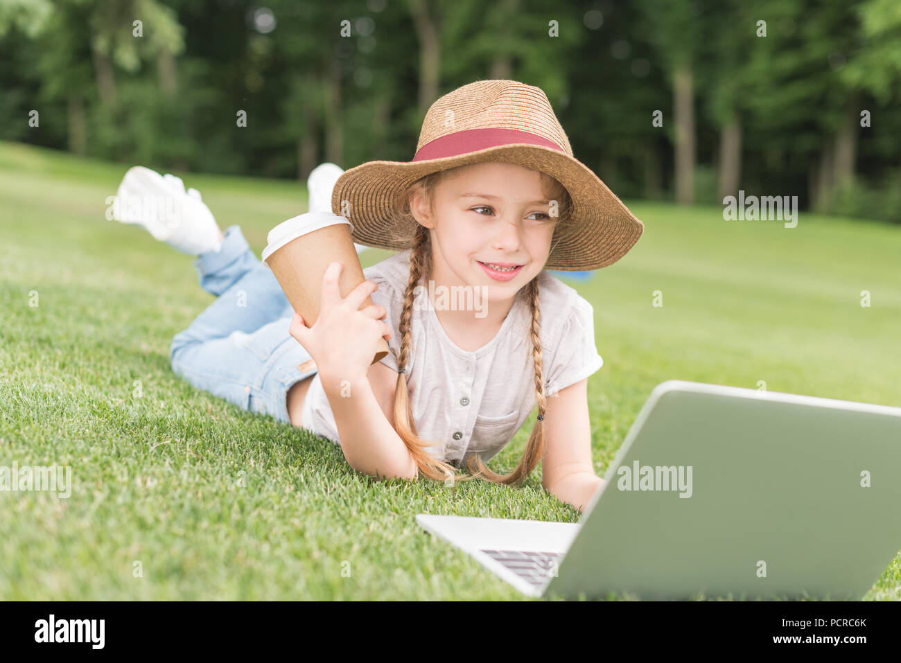 happy child holding paper cup and using laptop while lying on grass in park Stock Photo