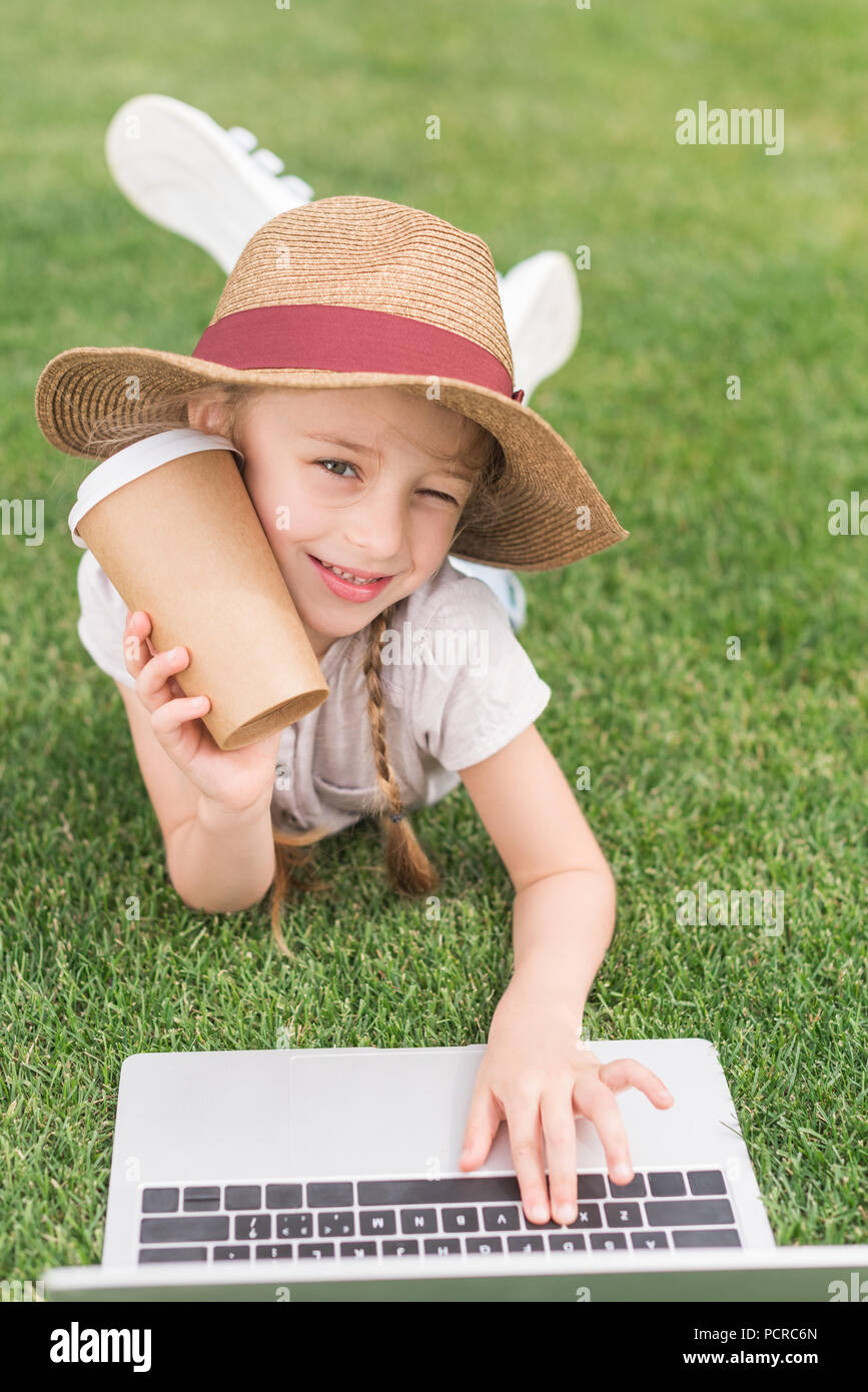 adorable child holding paper cup and smiling at camera while using laptop on green meadow Stock Photo