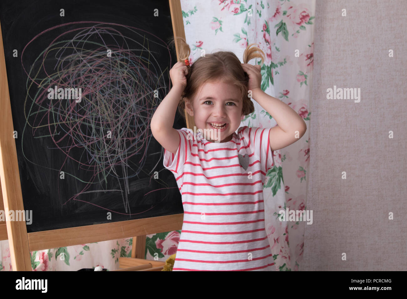 Child is drawing with pieces of color chalk on the chalk board. Girl is expressing creativity and looking at the camera. Concept of expression and learning Stock Photo
