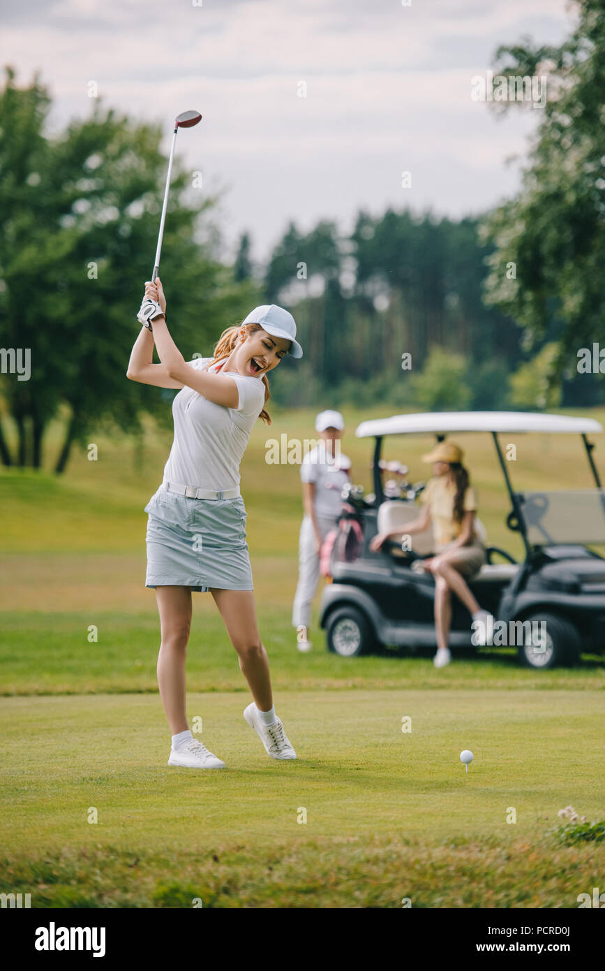 selective focus of woman with golf club playing golf and friends resting at golf cart on green lawn Stock Photo
