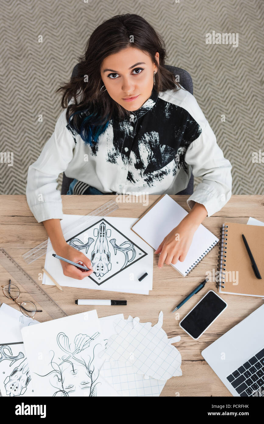overhead view of female artist painting and looking at camera while sitting at table with laptop and smartphone Stock Photo